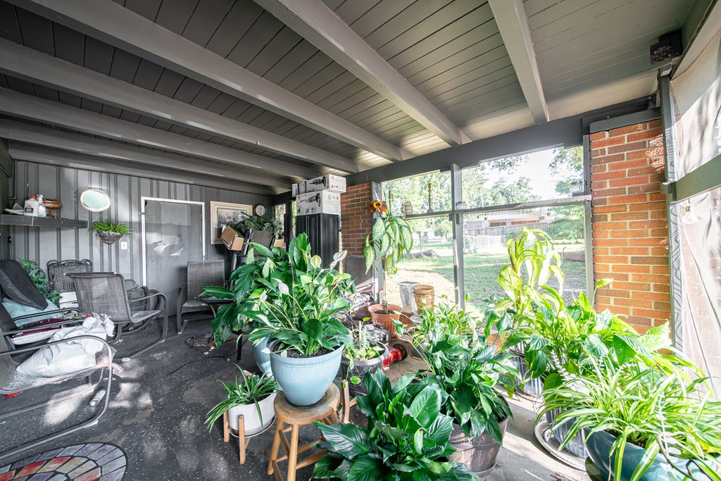 Enclosed sunroom with exposed beams, outdoor chairs, and abundant potted plants.