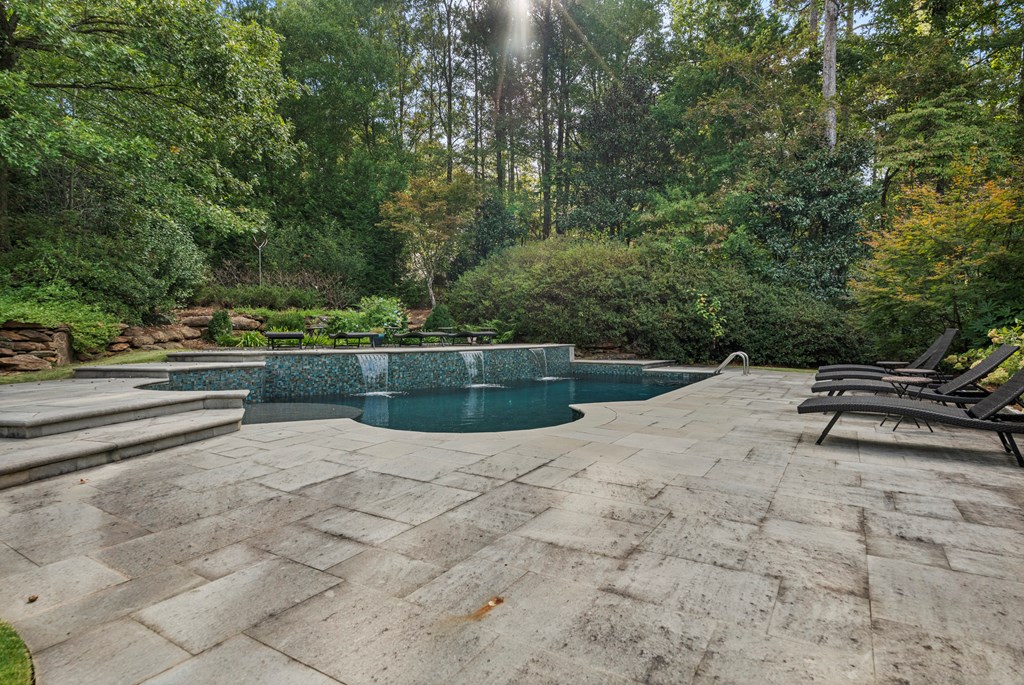 Swimming pool with tiled waterfall edge, stone patio, lounge chairs, and mature trees.