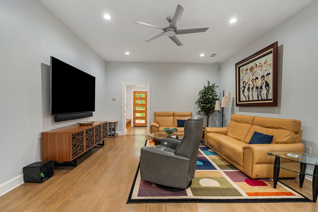 Staged living room with tan leather sofas, a gray recliner, colorful rug, and wall-mounted television.