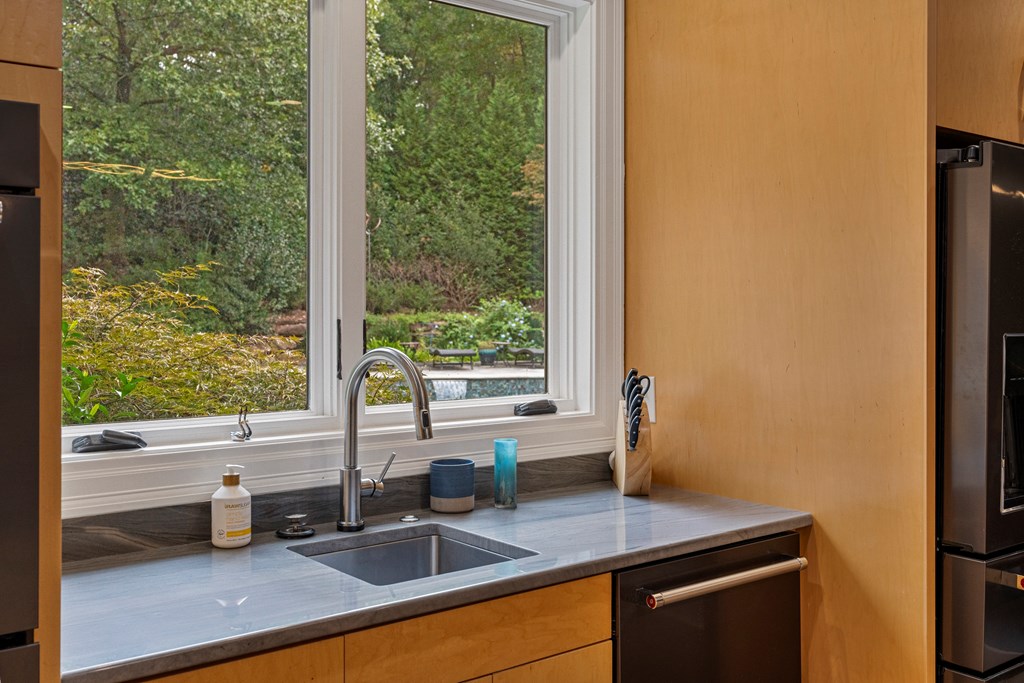 Kitchen sink beneath large windows overlooking a wooded yard and swimming pool.