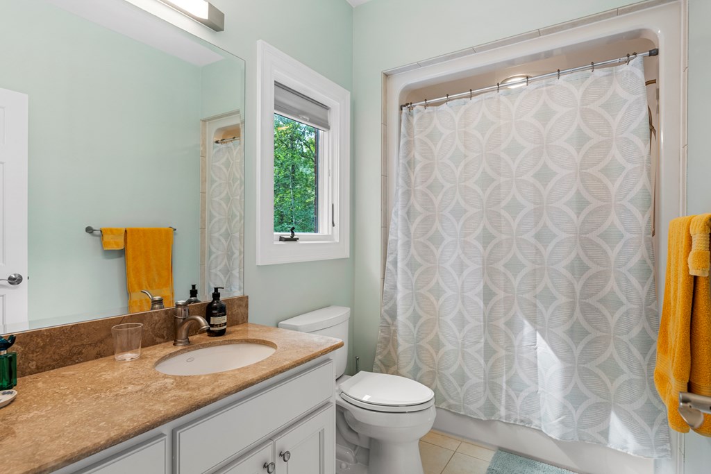 Bathroom with a tan stone vanity, patterned shower curtain, and window overlooking greenery.
