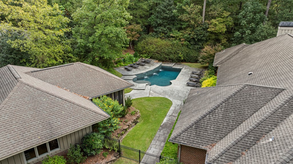 Aerial view of a backyard pool and paved patio surrounded by homes and dense trees.