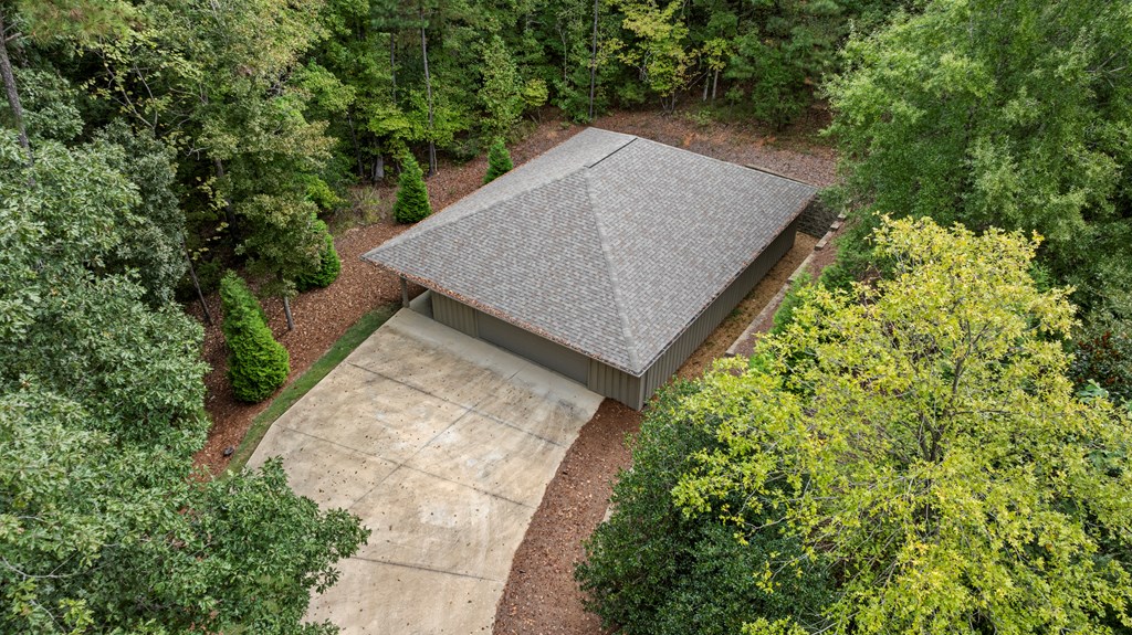 Aerial view of a low, shingle-roofed structure and concrete driveway surrounded by trees.