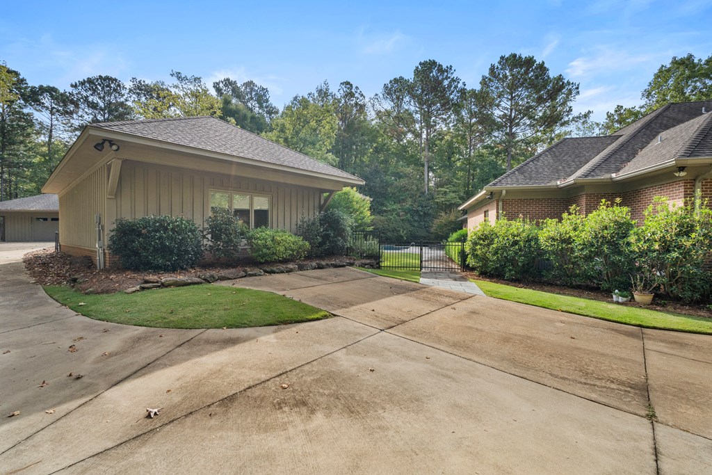 Concrete driveway and gated entrance between two buildings, bordered by shrubs and mature trees.
