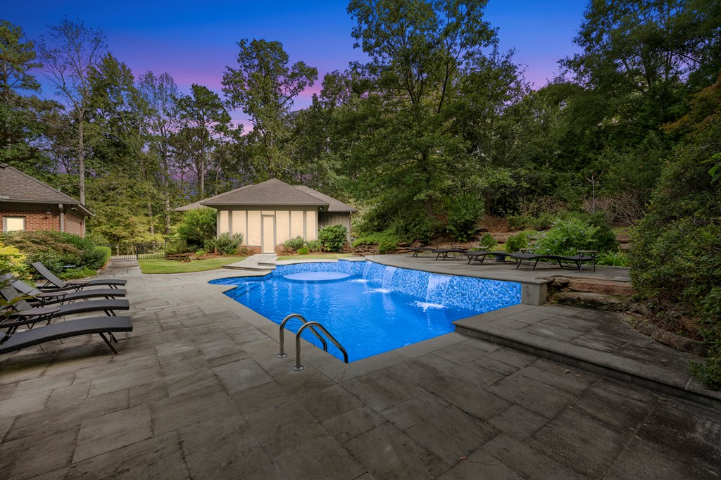 Swimming pool with a broad stone patio, lounge chairs, and a small pool house among mature trees.