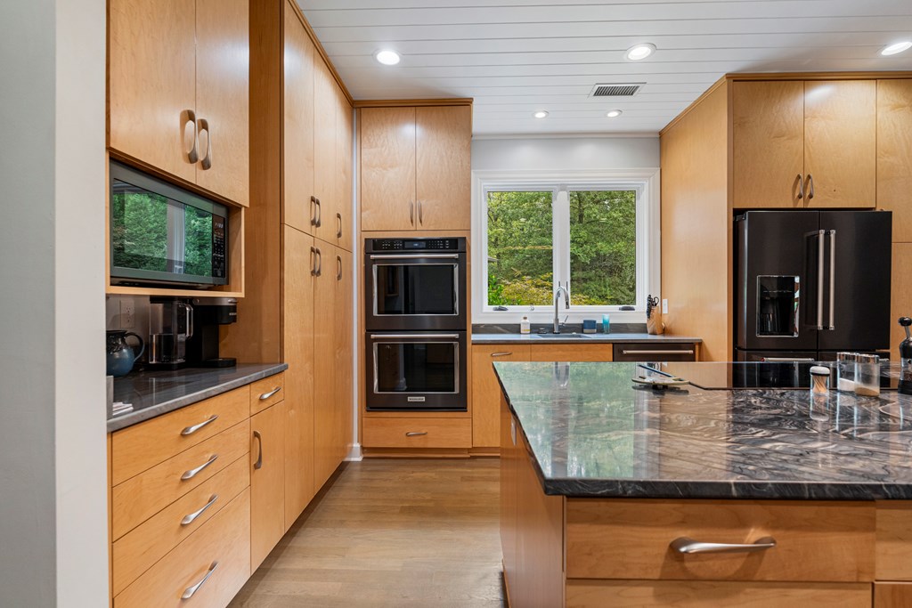 Kitchen with light wood cabinets, double ovens, and a large stone island beside a window overlooking trees.