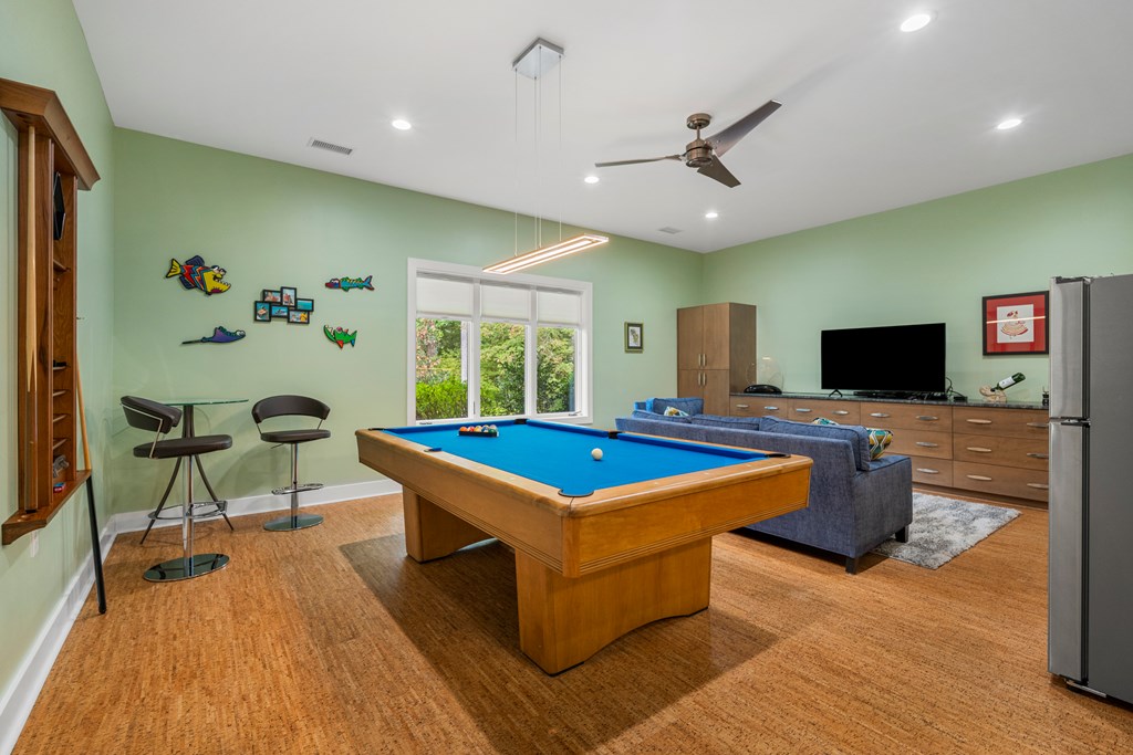 Game room with a blue-felt pool table, television seating area, and large window overlooking greenery.