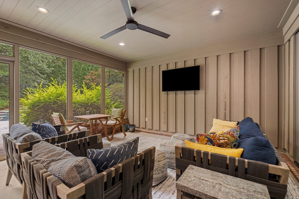Furnished enclosed porch with a ceiling fan, wall-mounted TV, and large windows overlooking greenery.
