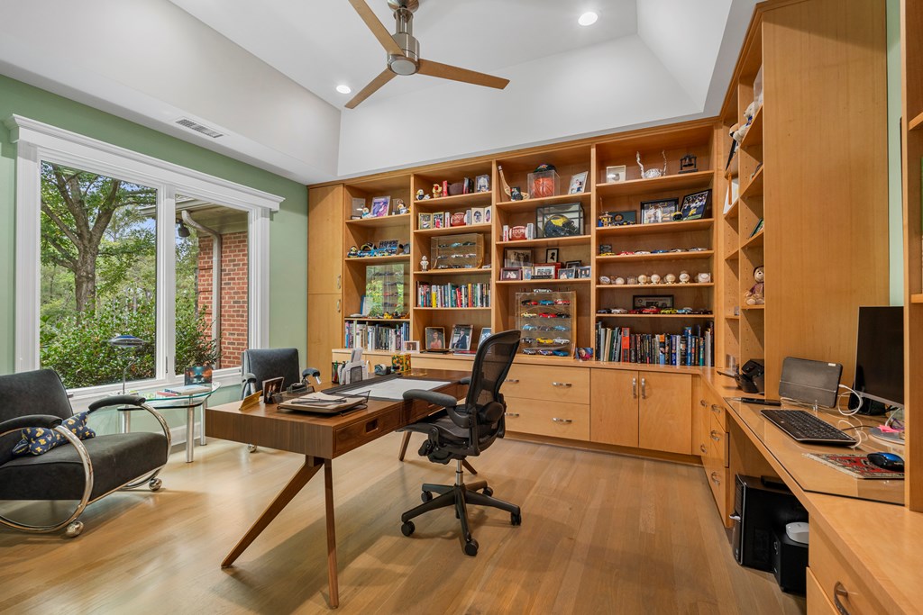 Home office with built-in wood shelving, two desks, and a large window overlooking greenery.