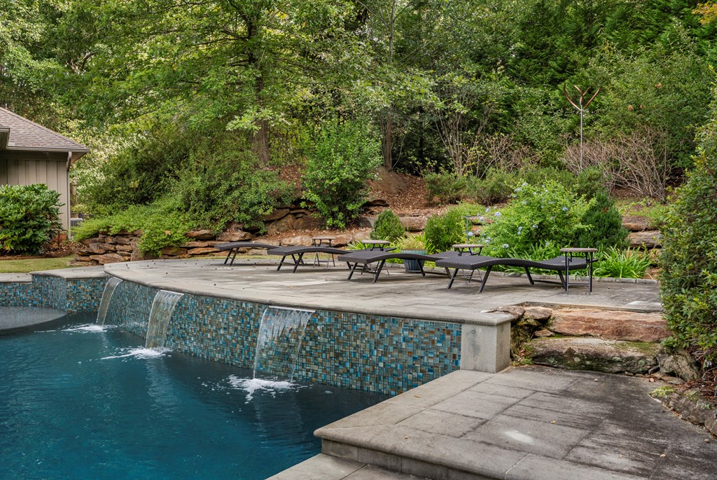 Swimming pool with tiled edge, cascading water, and lounge chairs on a stone patio.