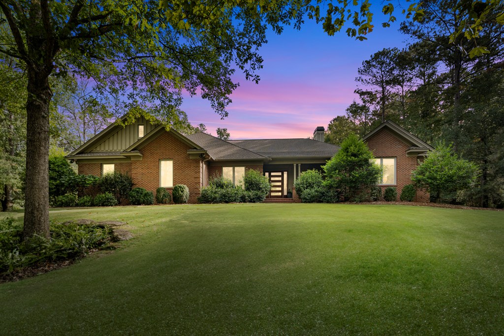 Brick home with illuminated windows and entry, framed by mature trees across a broad lawn at dusk.
