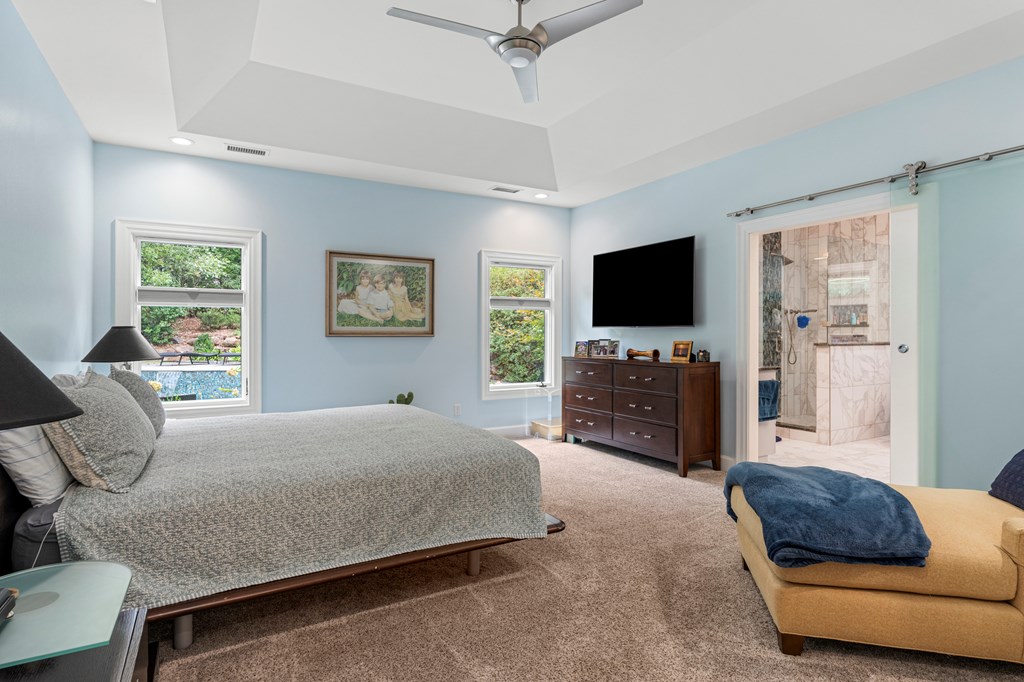 Furnished bedroom with pale blue walls, a tray ceiling, and a doorway to a marble-tiled bathroom.