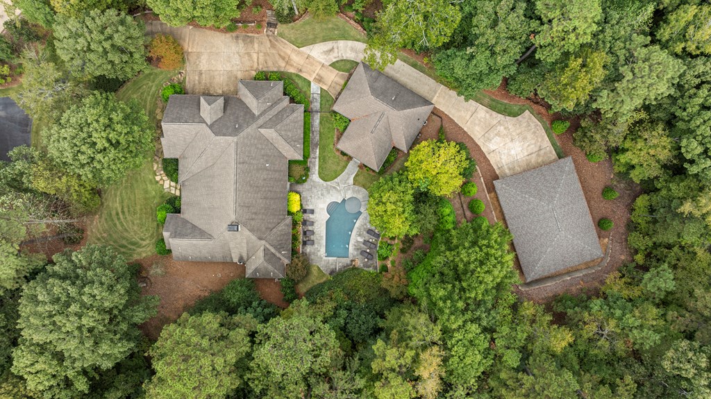 Aerial view of a wooded home with a swimming pool, outbuildings, and curved driveway.