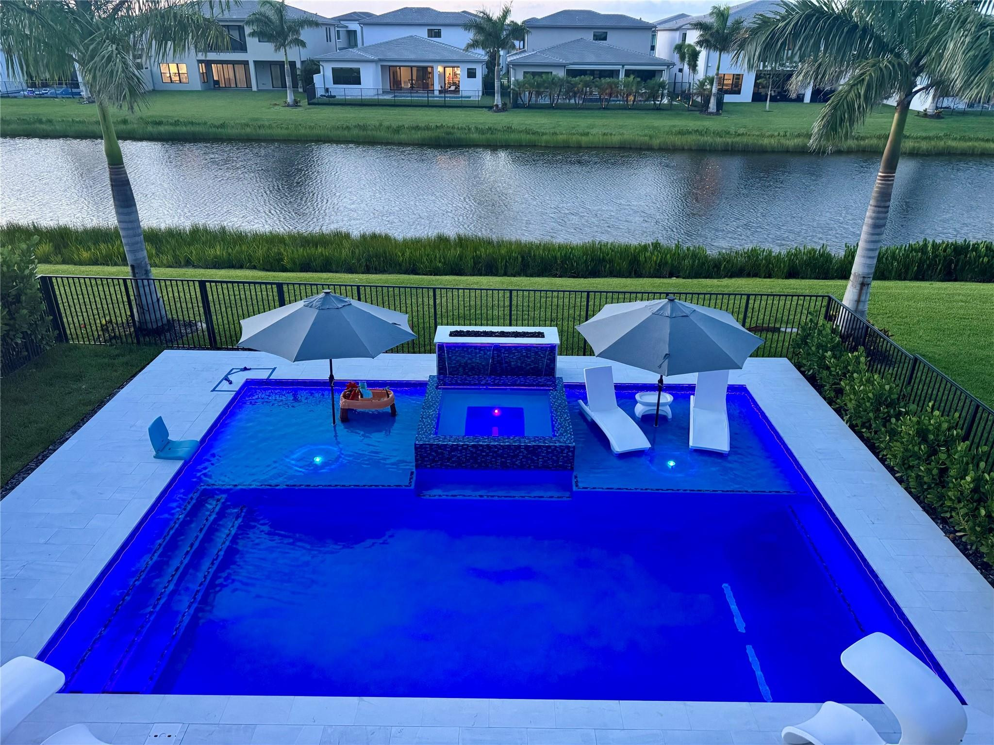 Blue-lit backyard pool and spa beside a canal, with lounge chairs and palm trees.