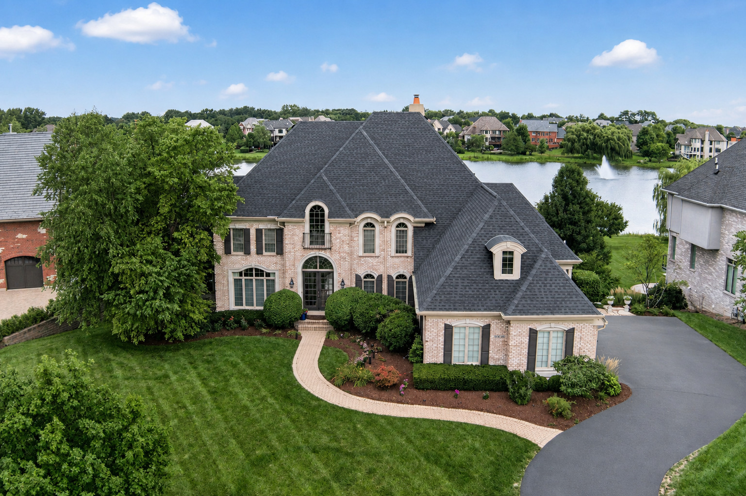 Aerial view of a brick house beside a lake, with a curved driveway and landscaped lawn.