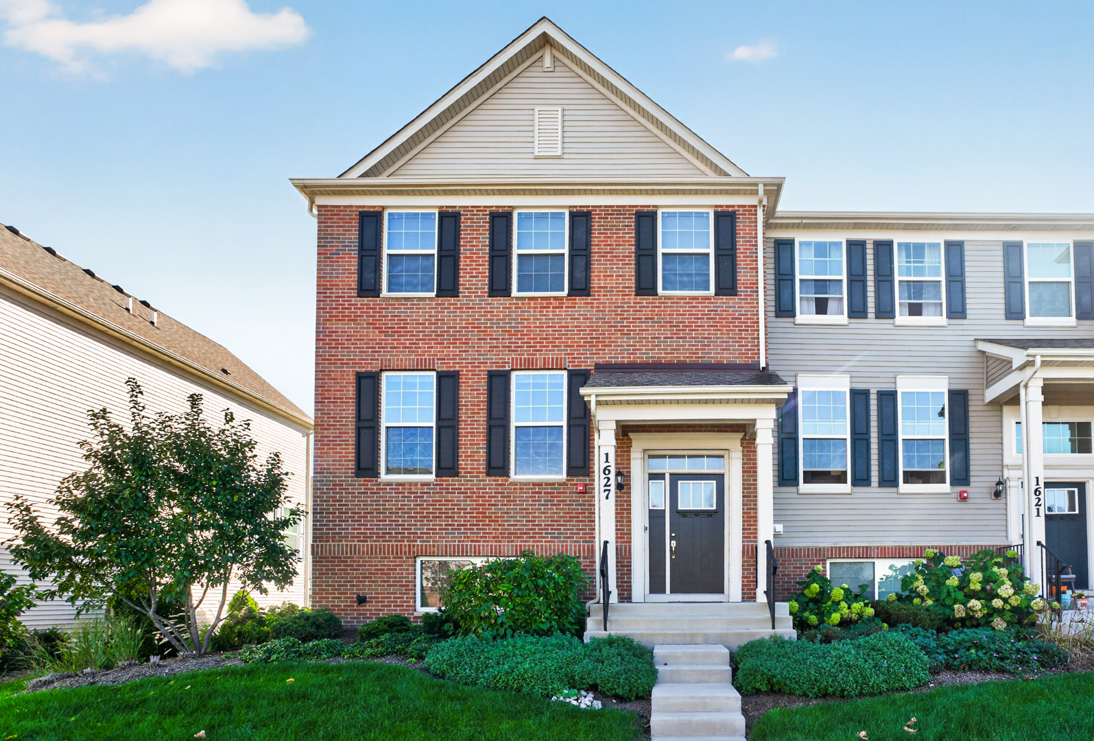 Red-brick townhouse with a white gable, dark shutters, covered entry, and landscaped front garden.