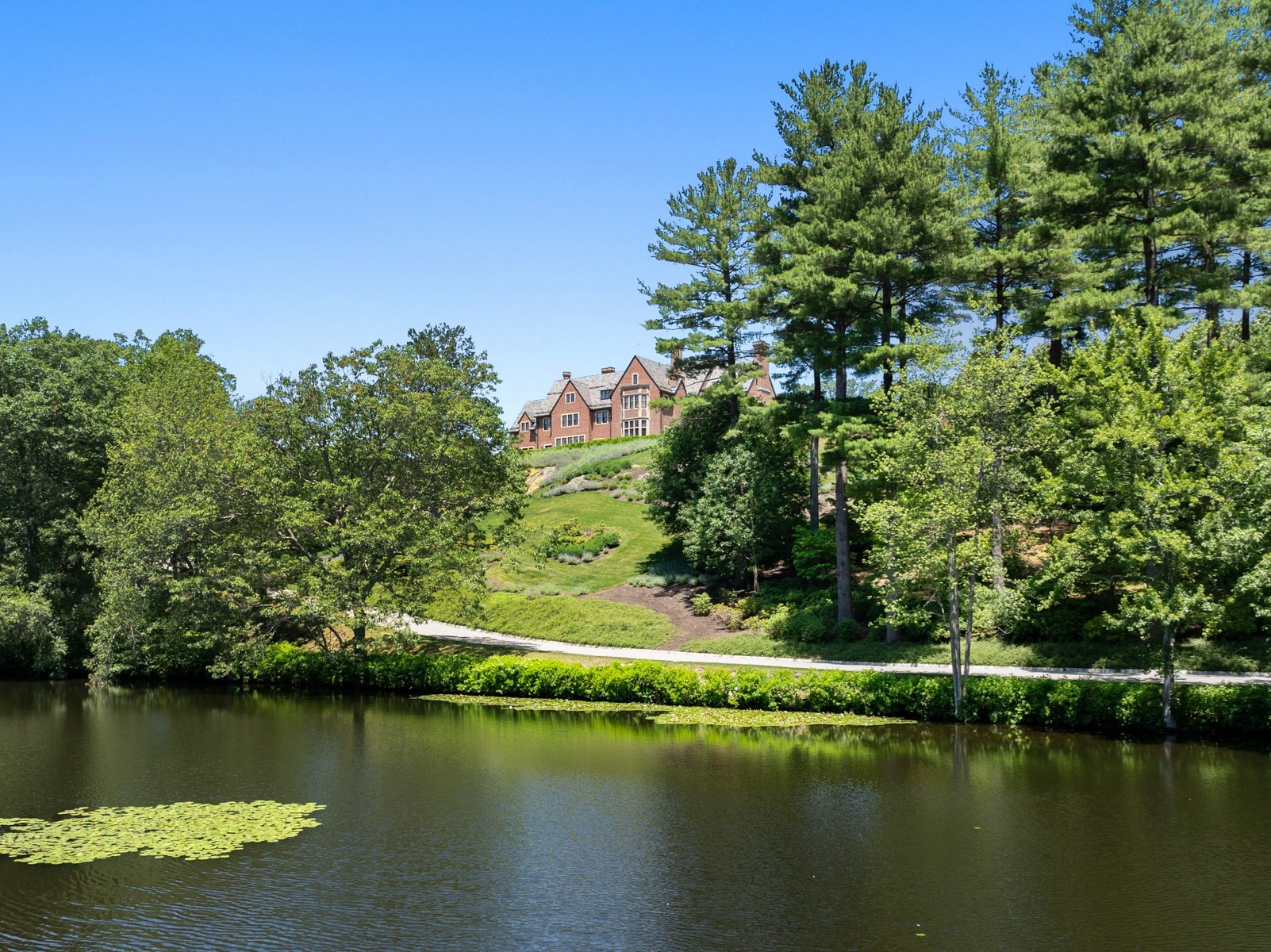 Red-brick house on a wooded hillside above a pond dotted with lily pads.