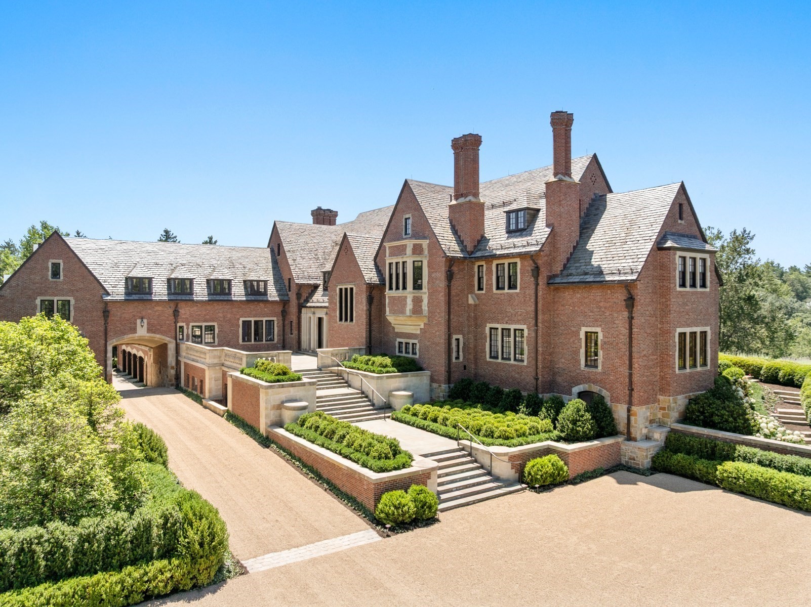 Red-brick residence with slate roofs and multiple chimneys, fronted by formal hedges and broad stone steps.