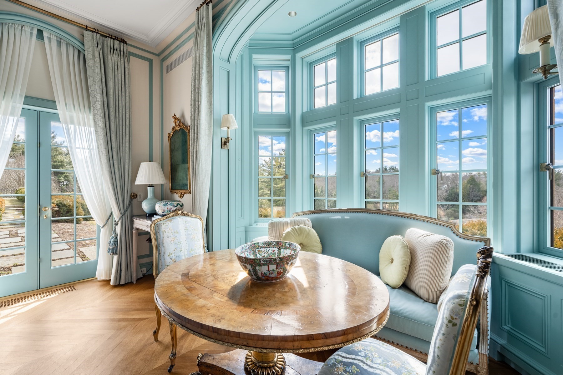 Turquoise sunroom with tall windows, curved sofa, and round wooden table.