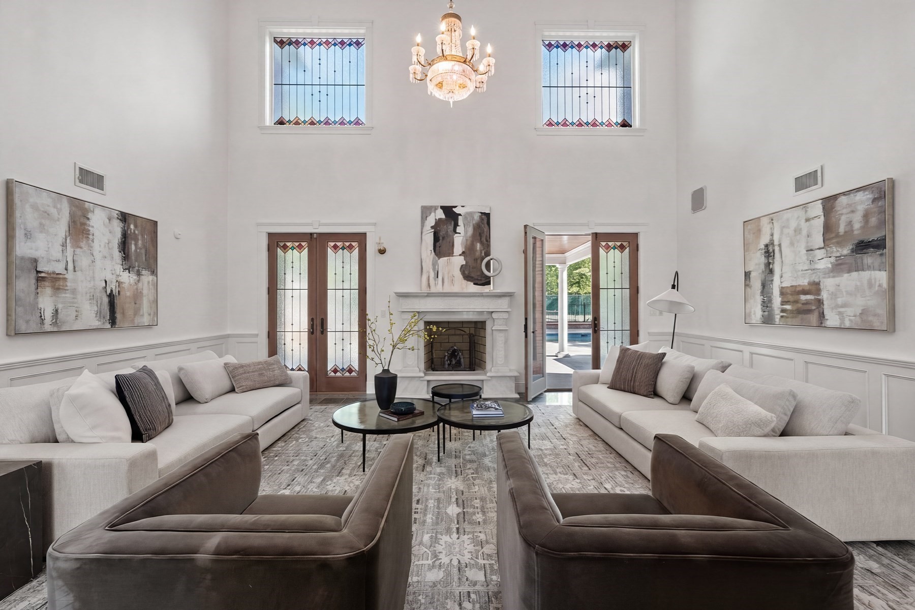 Double-height living room with stained-glass windows, a white fireplace, chandelier, and pale sofas.