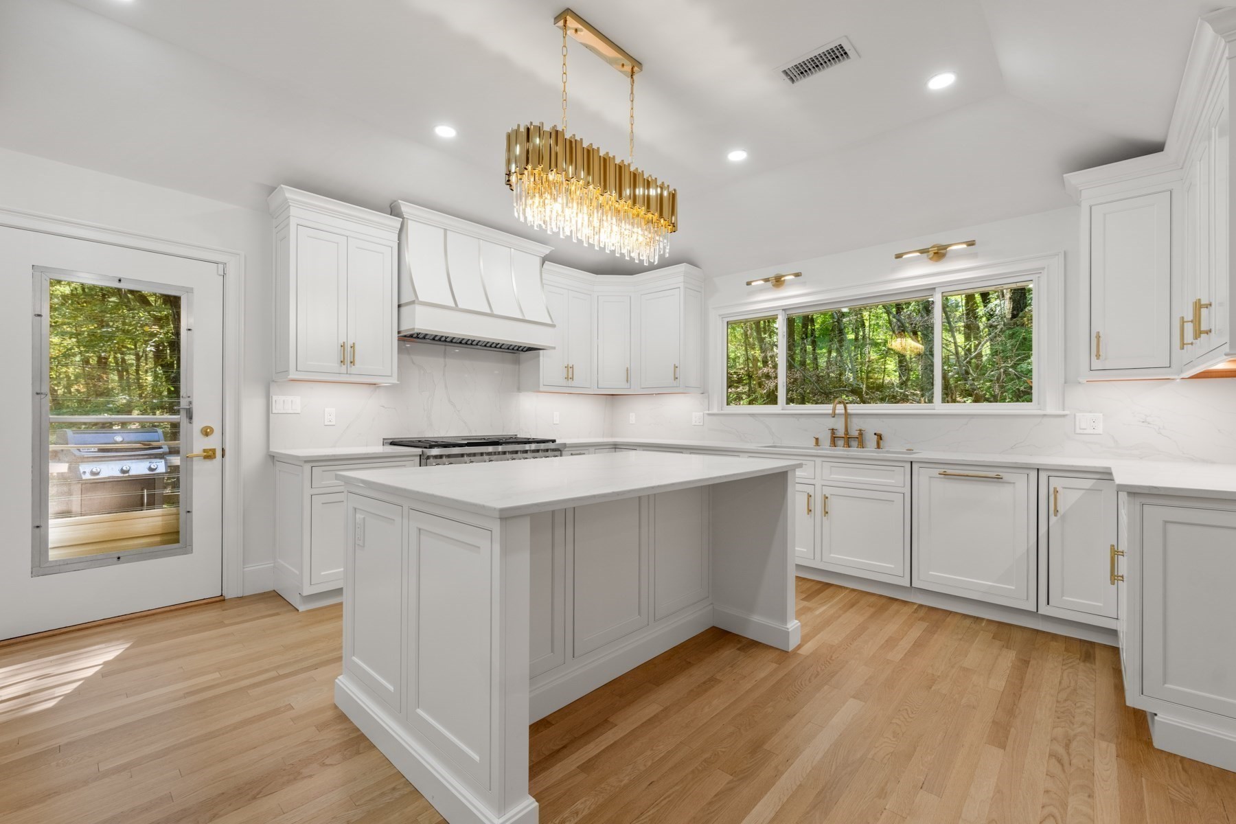 White kitchen with a large island, gold chandelier, and windows overlooking leafy woods.