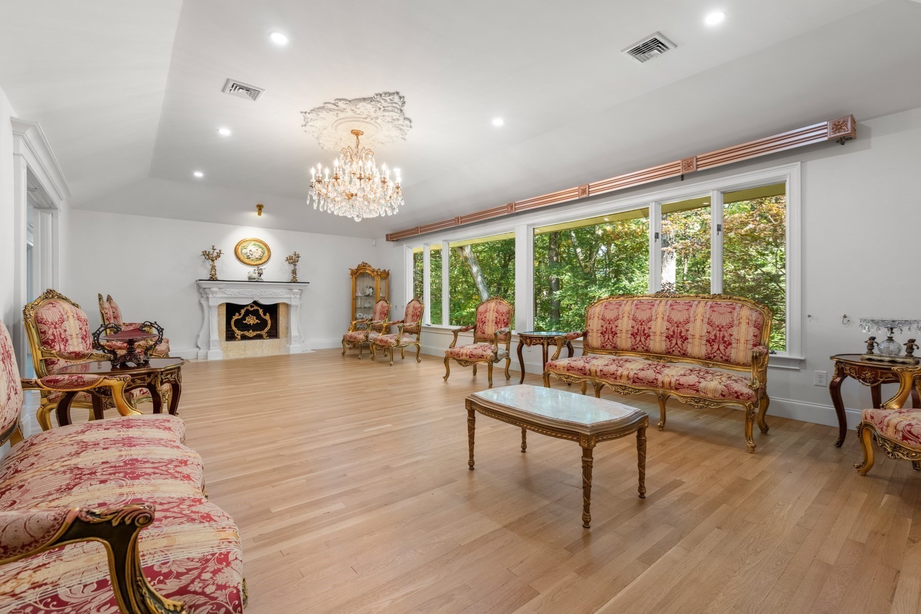 Living room with ornate red-and-gold furniture, a white fireplace, and large windows overlooking trees.