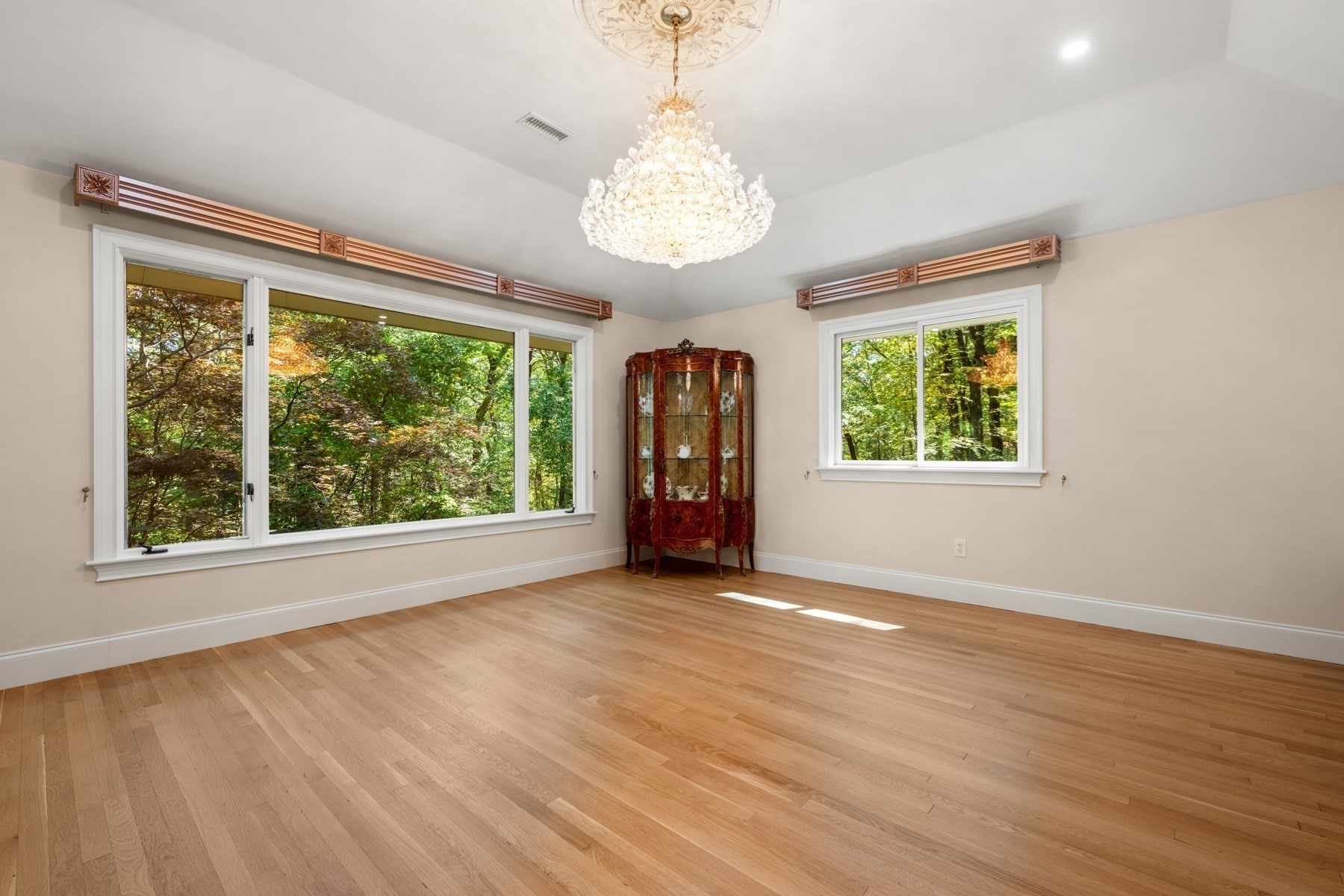 Vacant room with hardwood floors, crystal chandelier, and large windows overlooking trees.