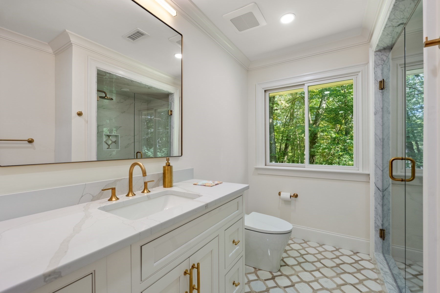 Bathroom with a marble vanity, brass fixtures, glass shower, patterned tile floor, and wooded view.