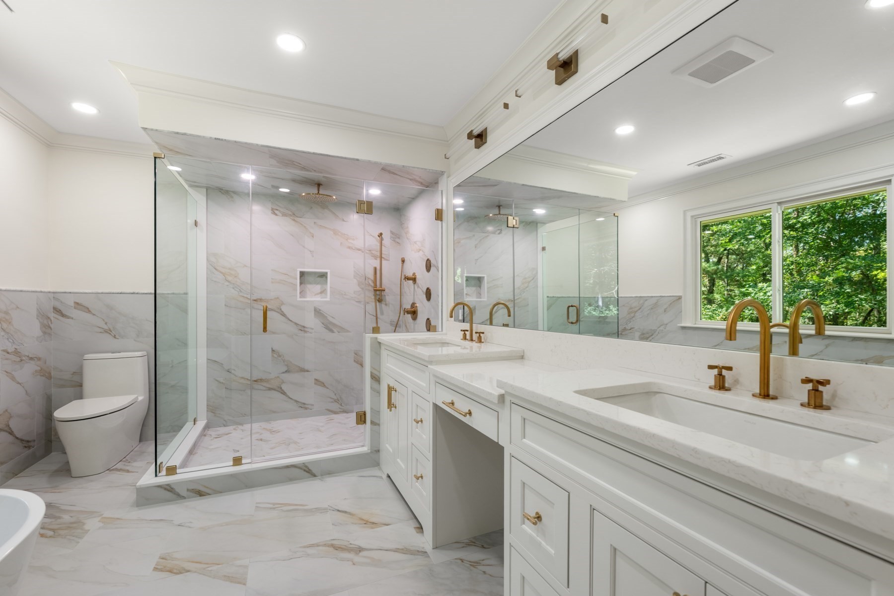 Bathroom with a glass walk-in shower, marble-look tile, double vanity, and a window overlooking trees.