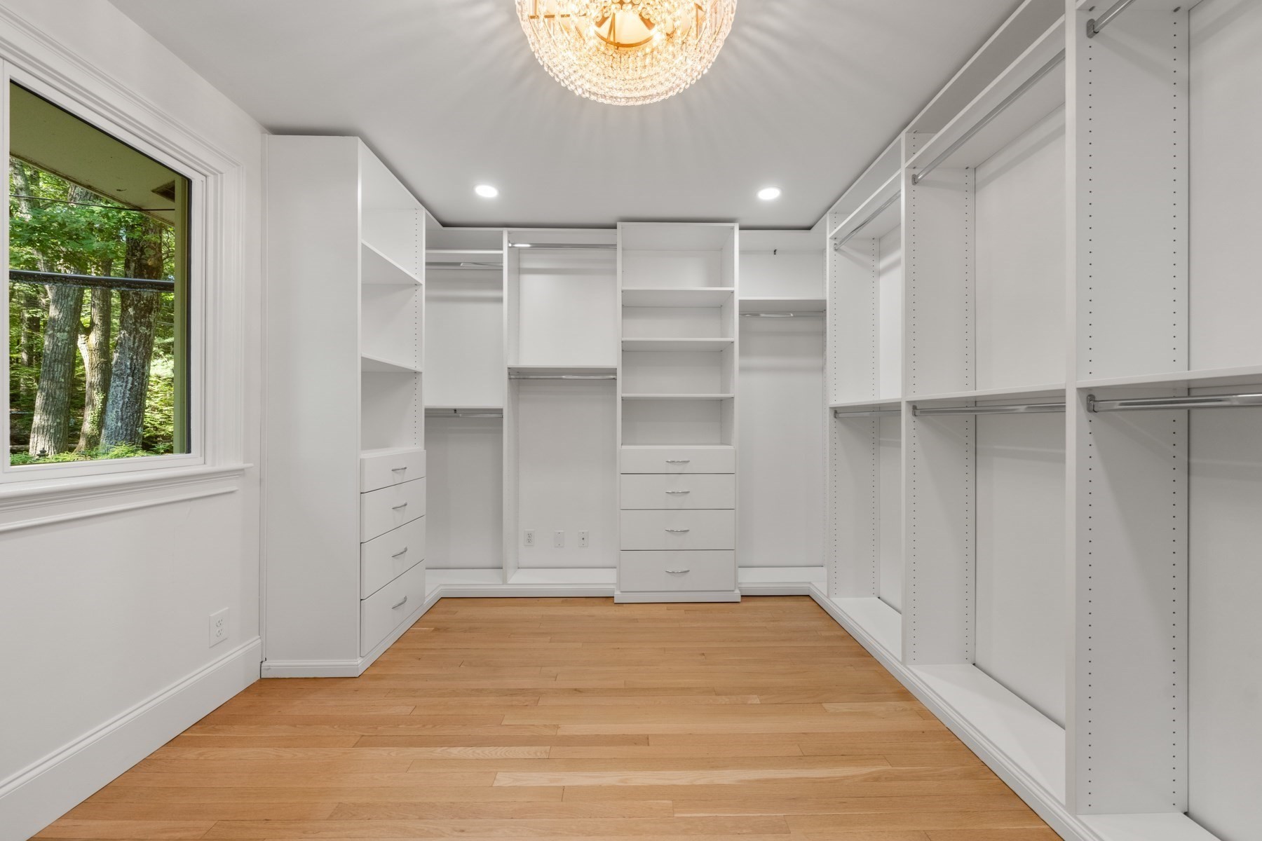 Walk-in closet with white built-ins, a crystal chandelier, hardwood flooring, and a window overlooking trees.