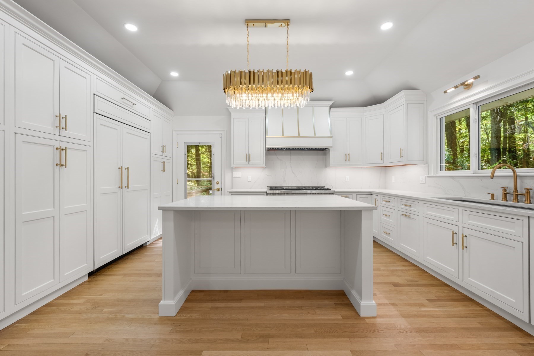 Bright white kitchen with a large island, gold chandelier, and windows overlooking trees.