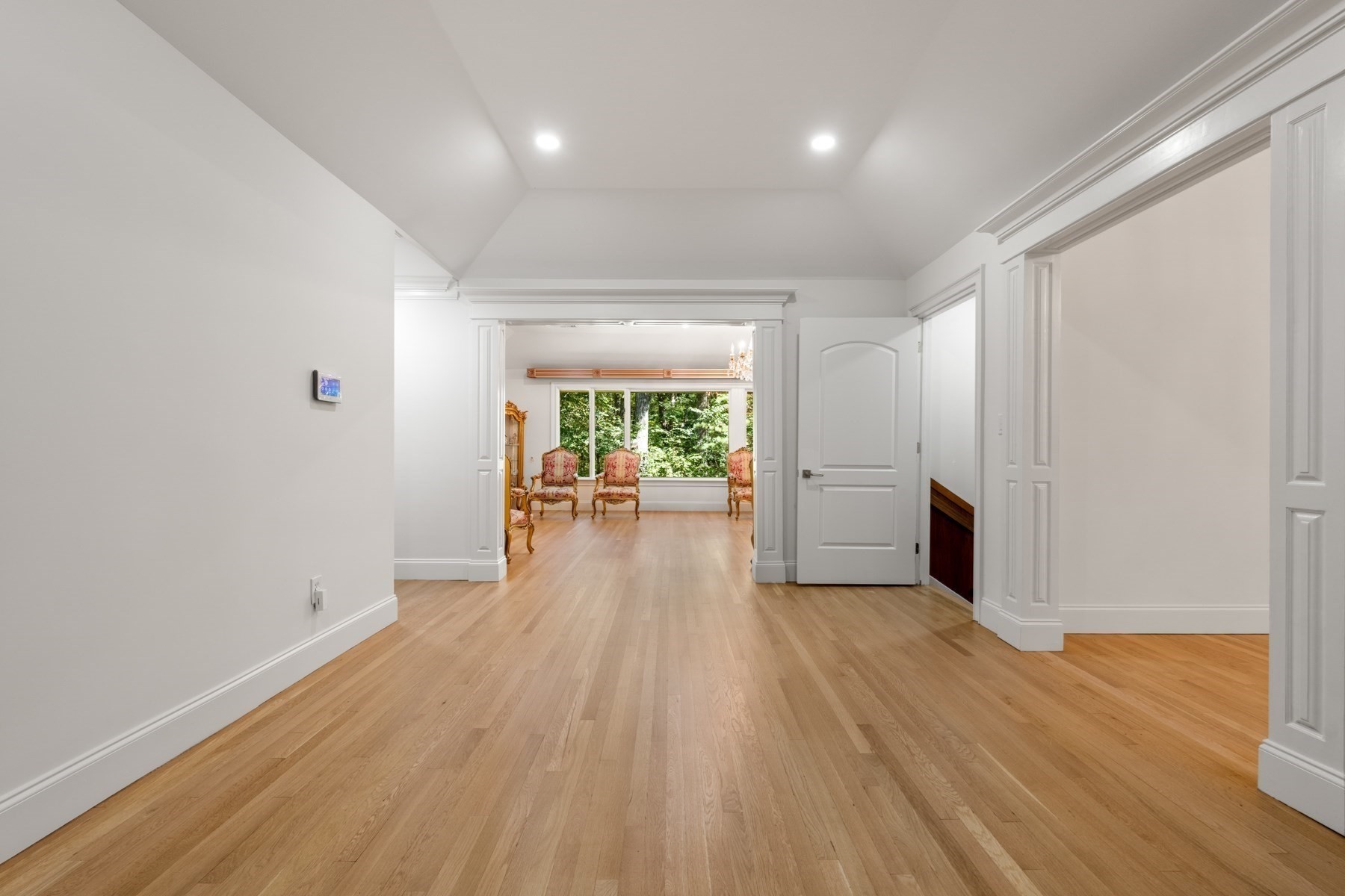 Hallway with light hardwood floors opens to a sitting room with upholstered chairs and a large window.
