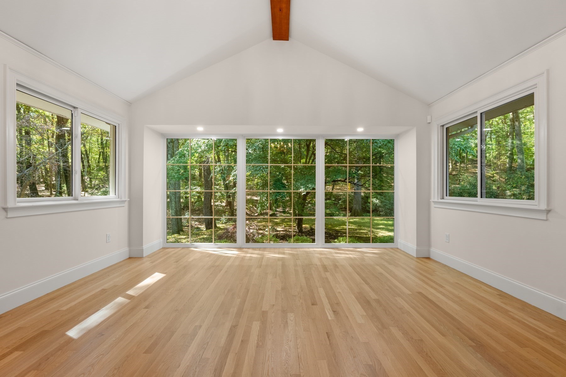 Empty vaulted room with hardwood floors and large windows overlooking trees.