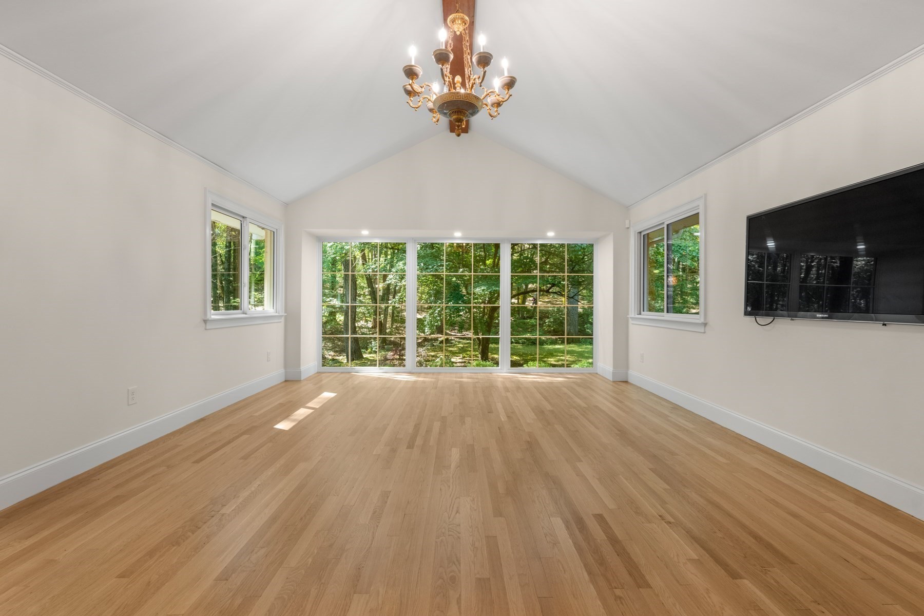 Bright empty room with vaulted ceiling, hardwood floors, large windows overlooking trees, and a chandelier.