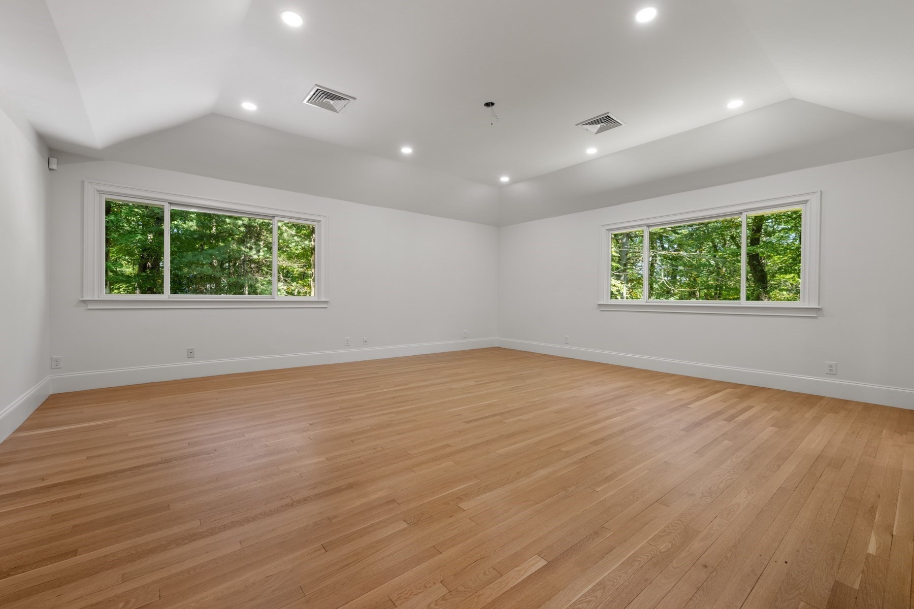 Empty room with pale hardwood floors and wide windows overlooking leafy trees.
