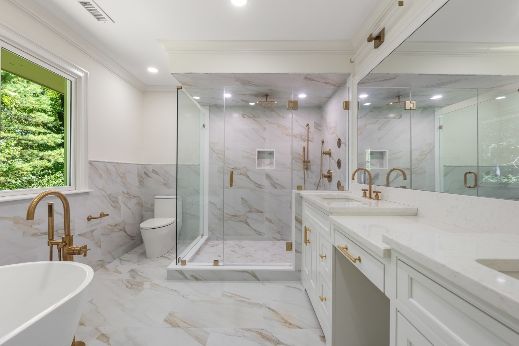 Marble bathroom with a glass shower, brass fixtures, double vanity, and freestanding tub beside a leafy window.