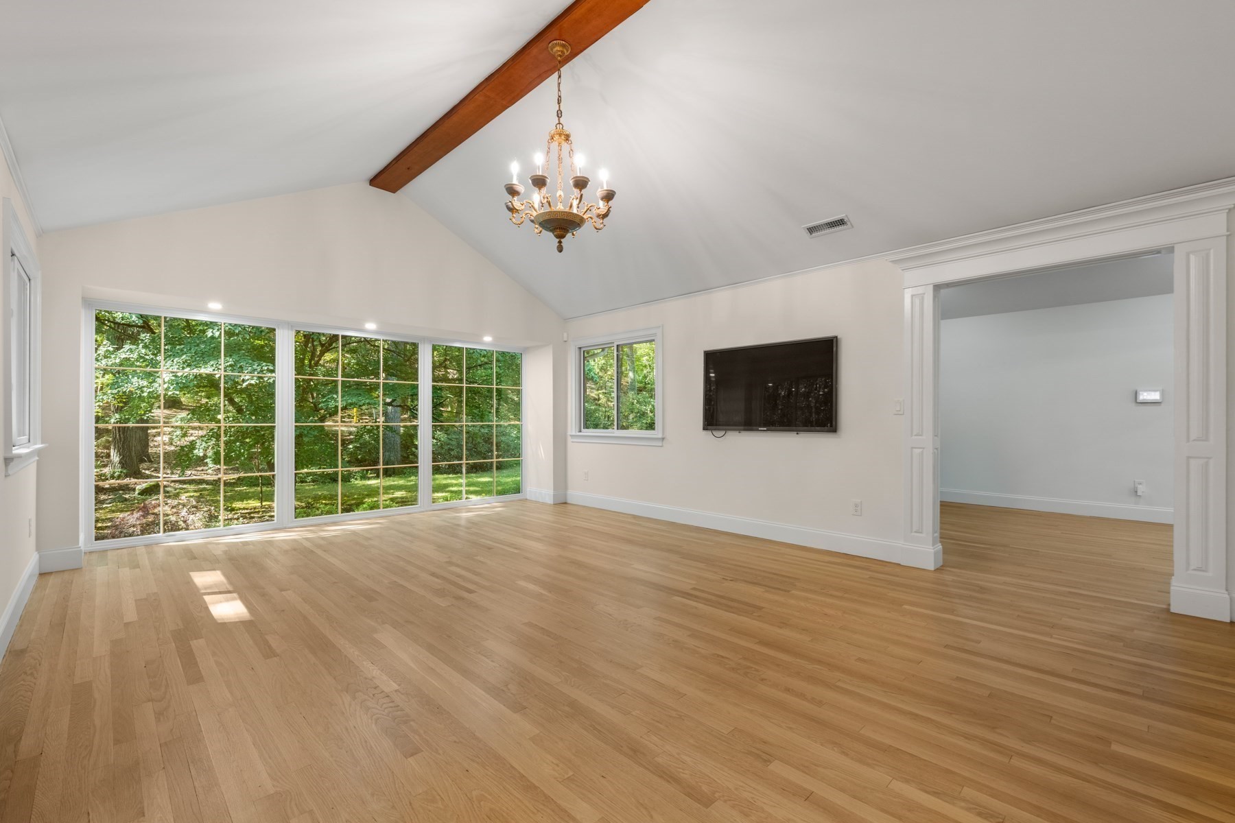 Vacant living room with vaulted ceiling, exposed beam, hardwood floors, and large windows overlooking trees.
