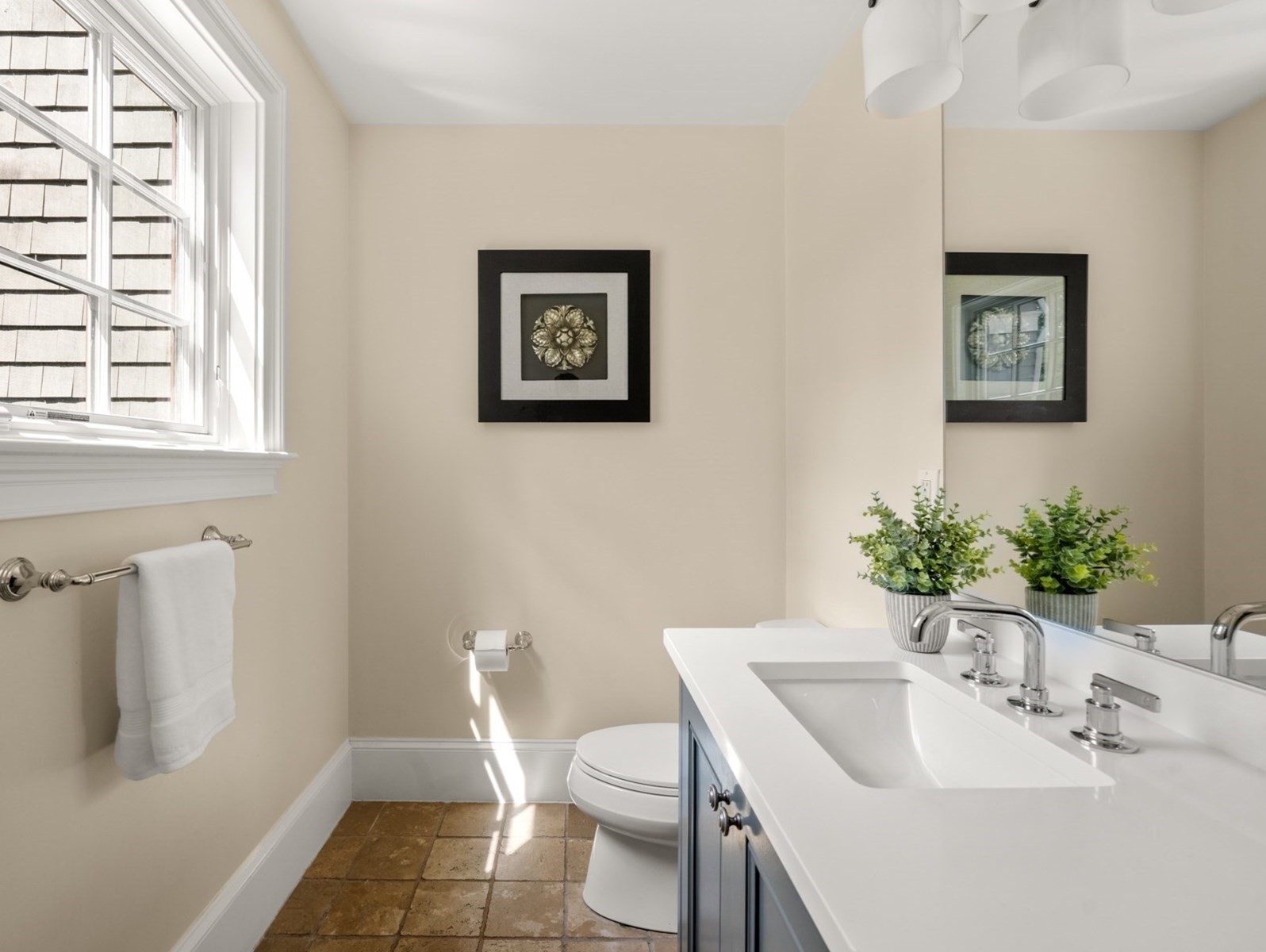 Bright bathroom with a white vanity, framed floral artwork, a window, and tan tiled floor.