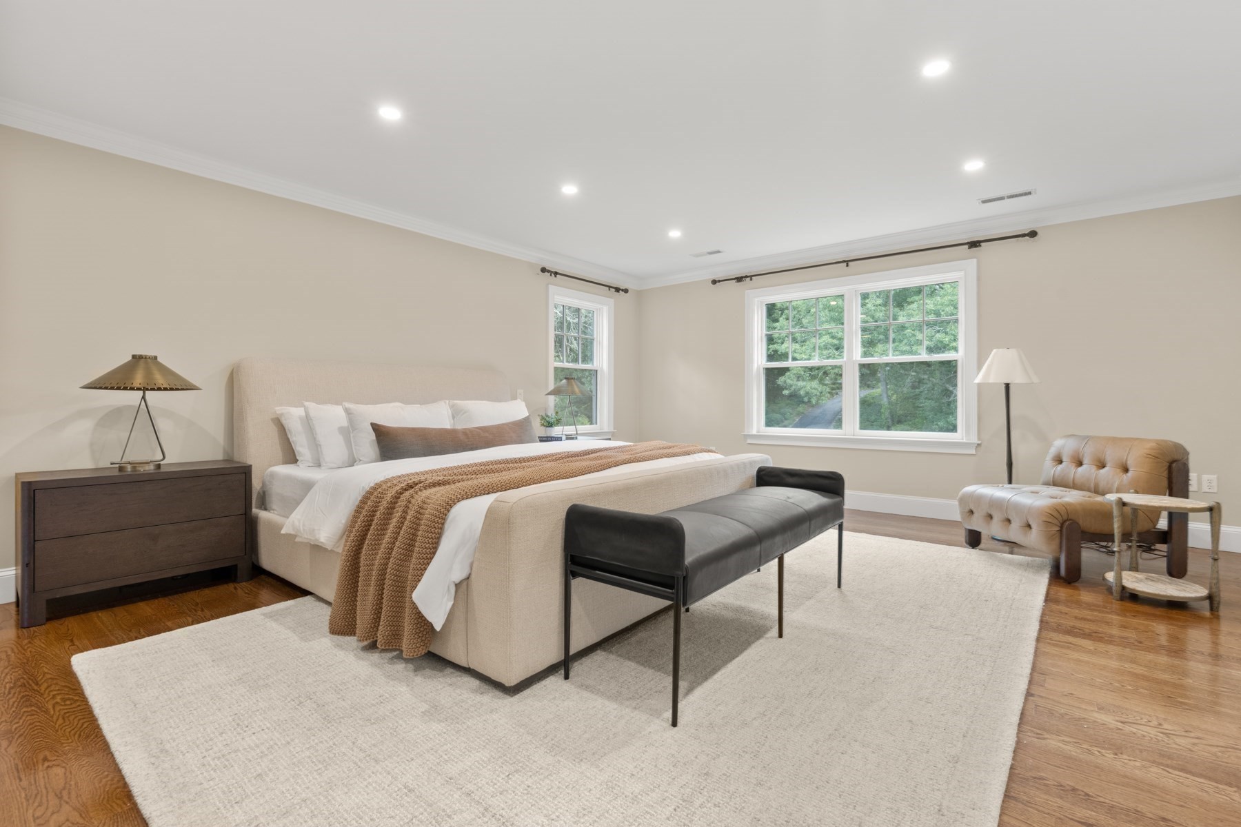 Staged bedroom with a king bed, dark bench, leather armchair, and windows overlooking trees.