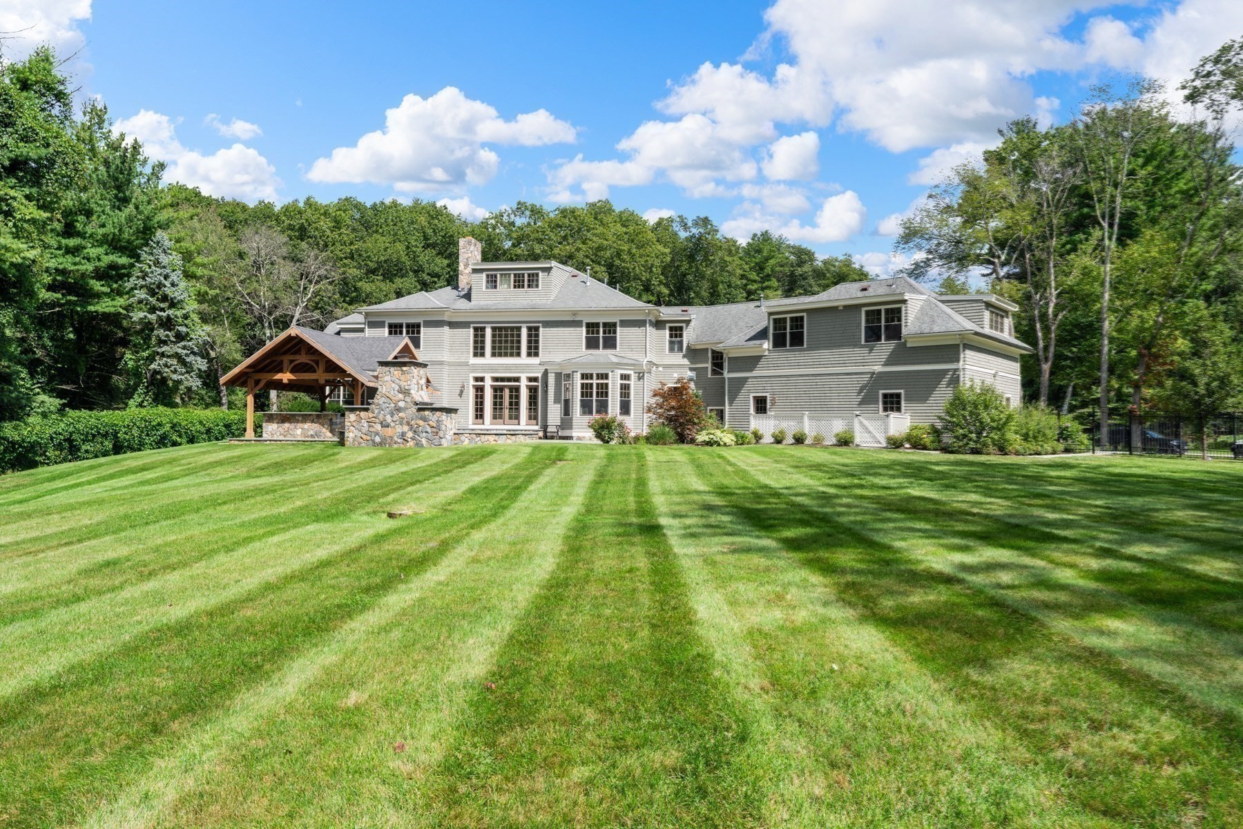 Rear of a large gray-shingled home with a broad lawn and stone-and-timber pavilion.