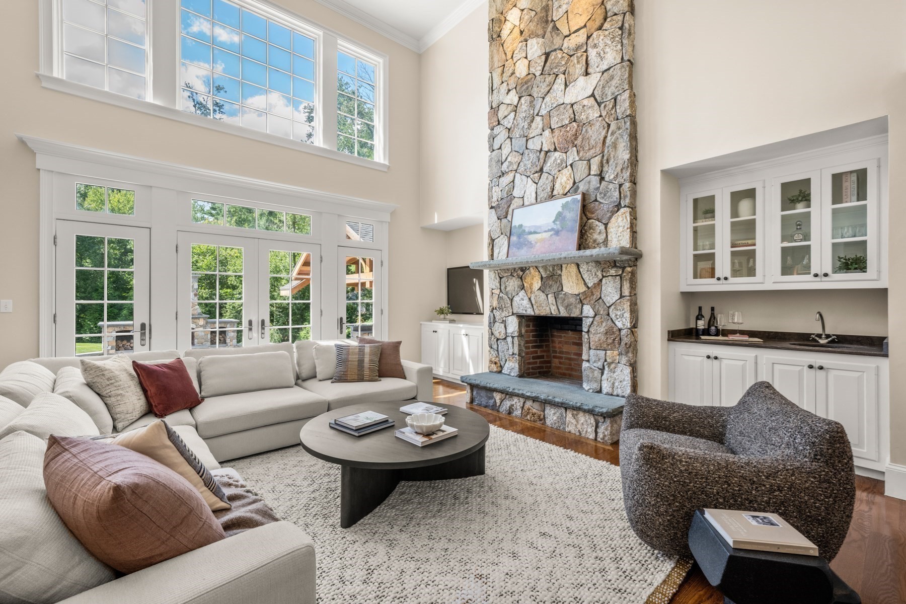 Living room with a tall stone fireplace, white sectional, high windows and built-in wet bar.