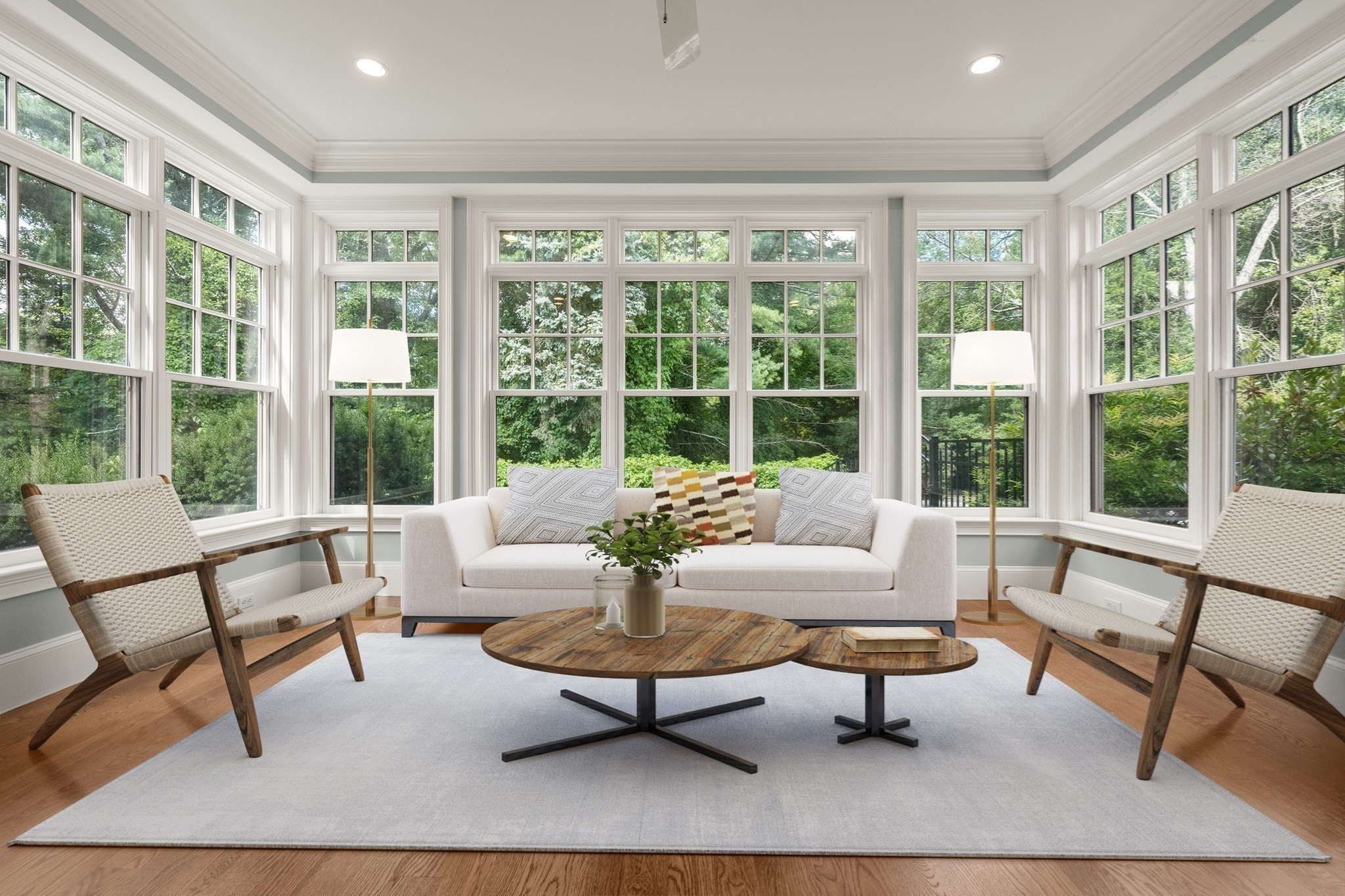 Sitting room with a white sofa and woven chairs surrounded by large windows overlooking trees.