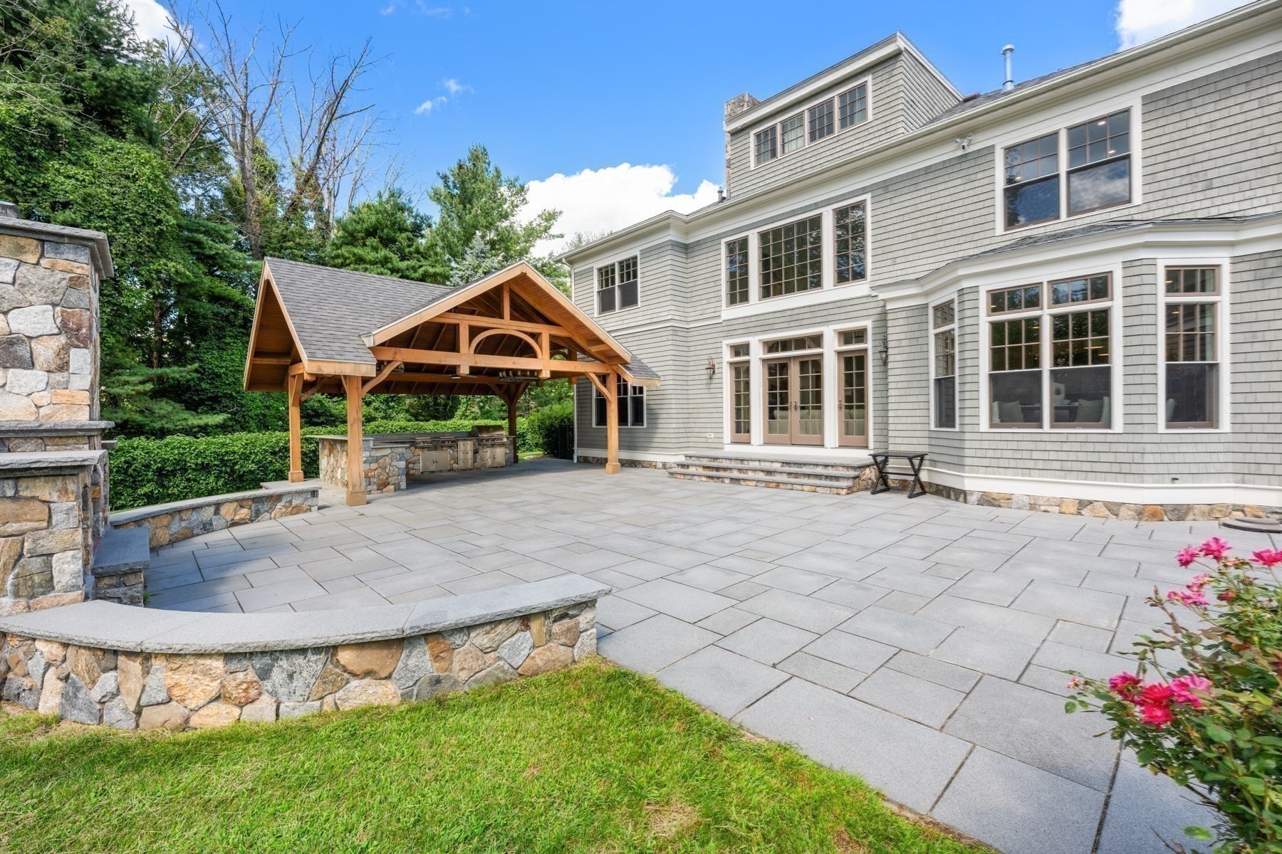 Gray-shingle home with a stone patio, timber pavilion, and outdoor kitchen.