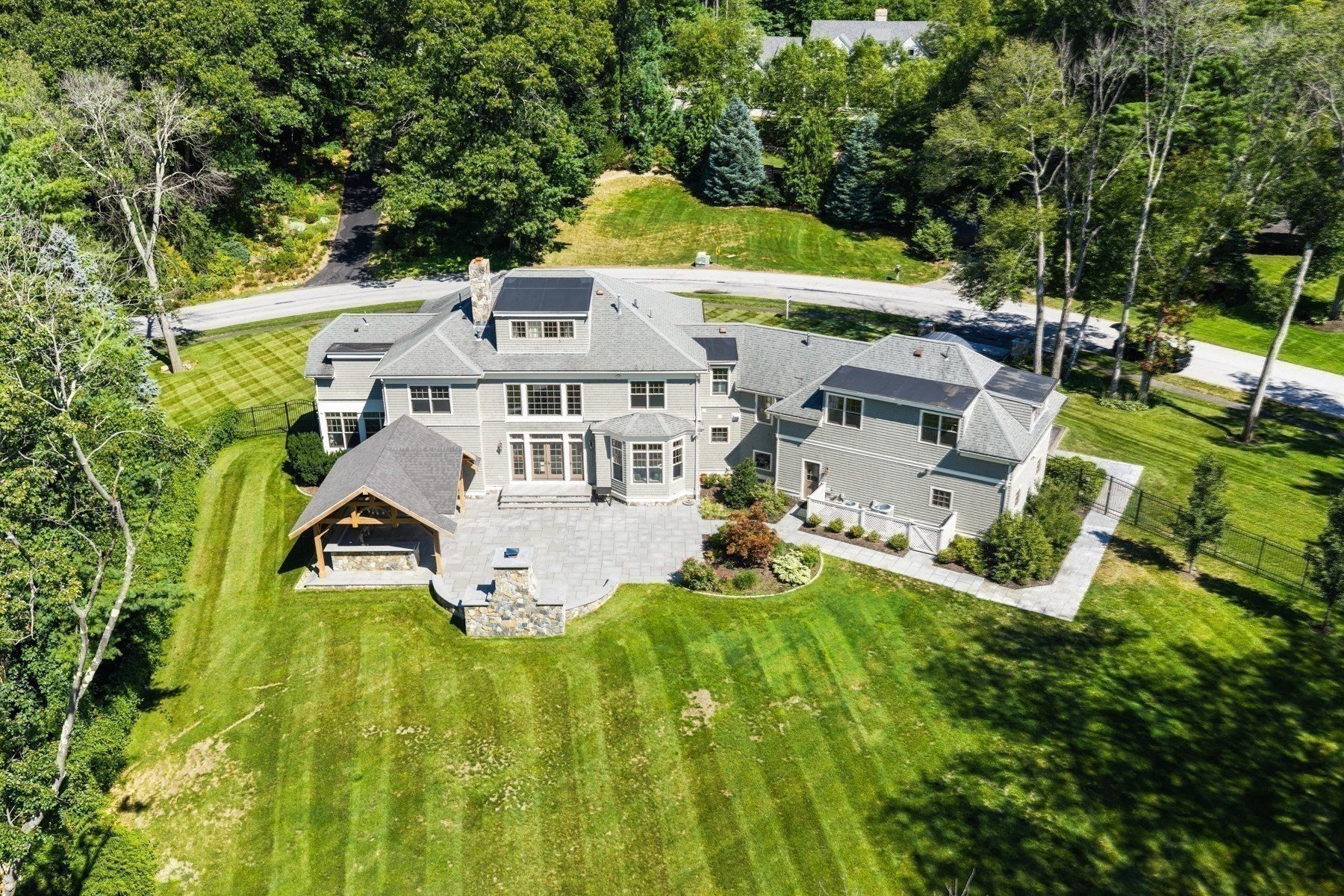 Aerial view of a large gray-shingled home with a stone patio and covered fireplace pavilion.