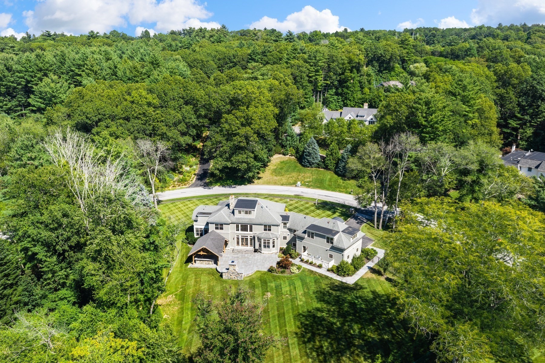 Aerial view of a gray house surrounded by woodland, with a broad lawn and stone patio.