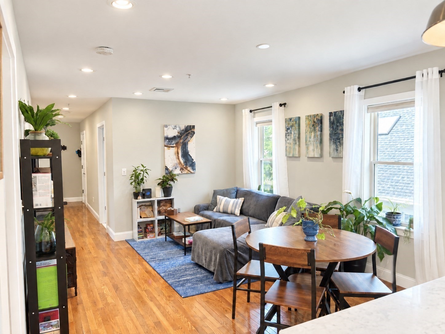 Open-plan living and dining area with hardwood floors, gray sofa, round wooden table, and large windows.