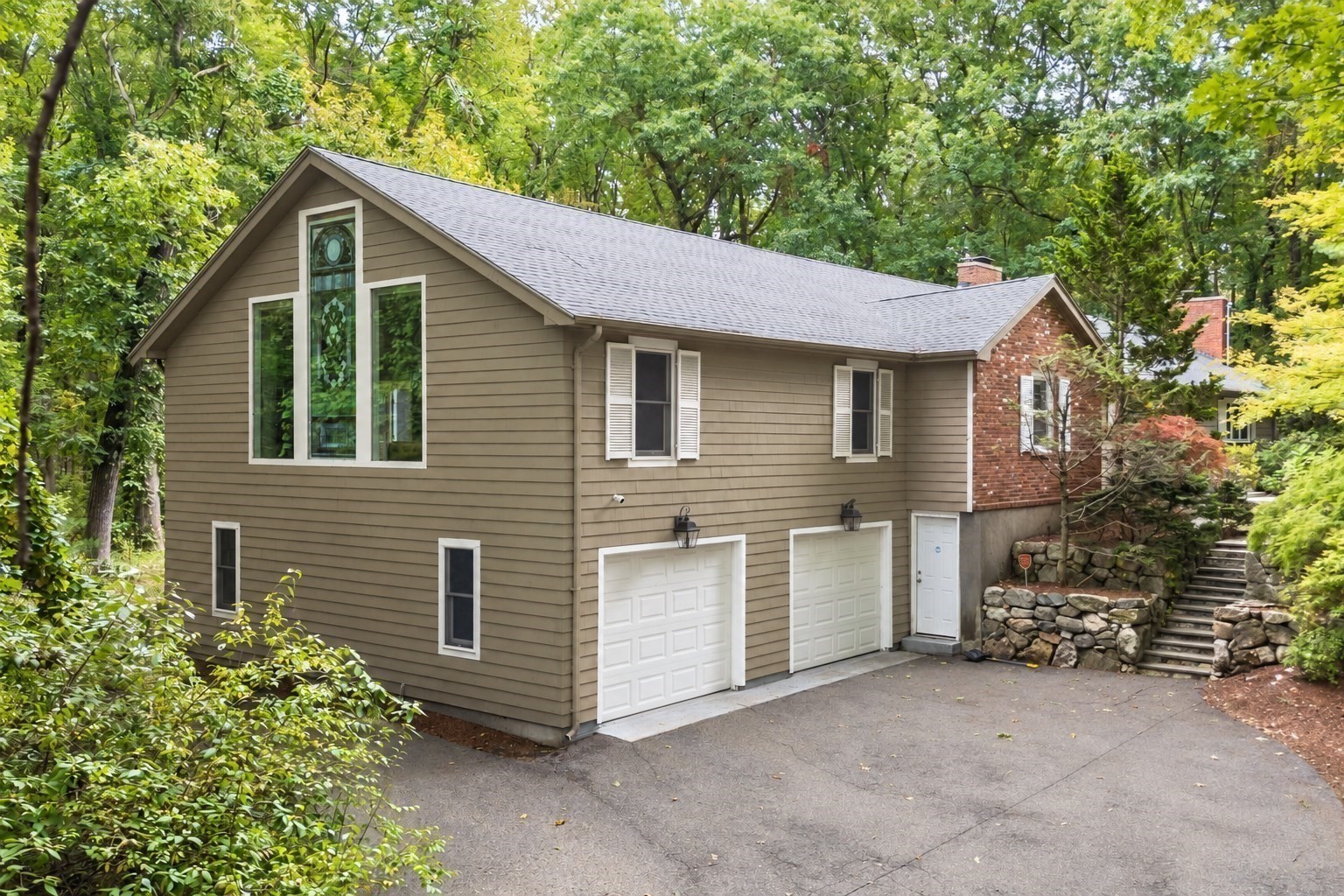 Taupe-sided home with two garage doors, a tall peaked window, and a wooded driveway.