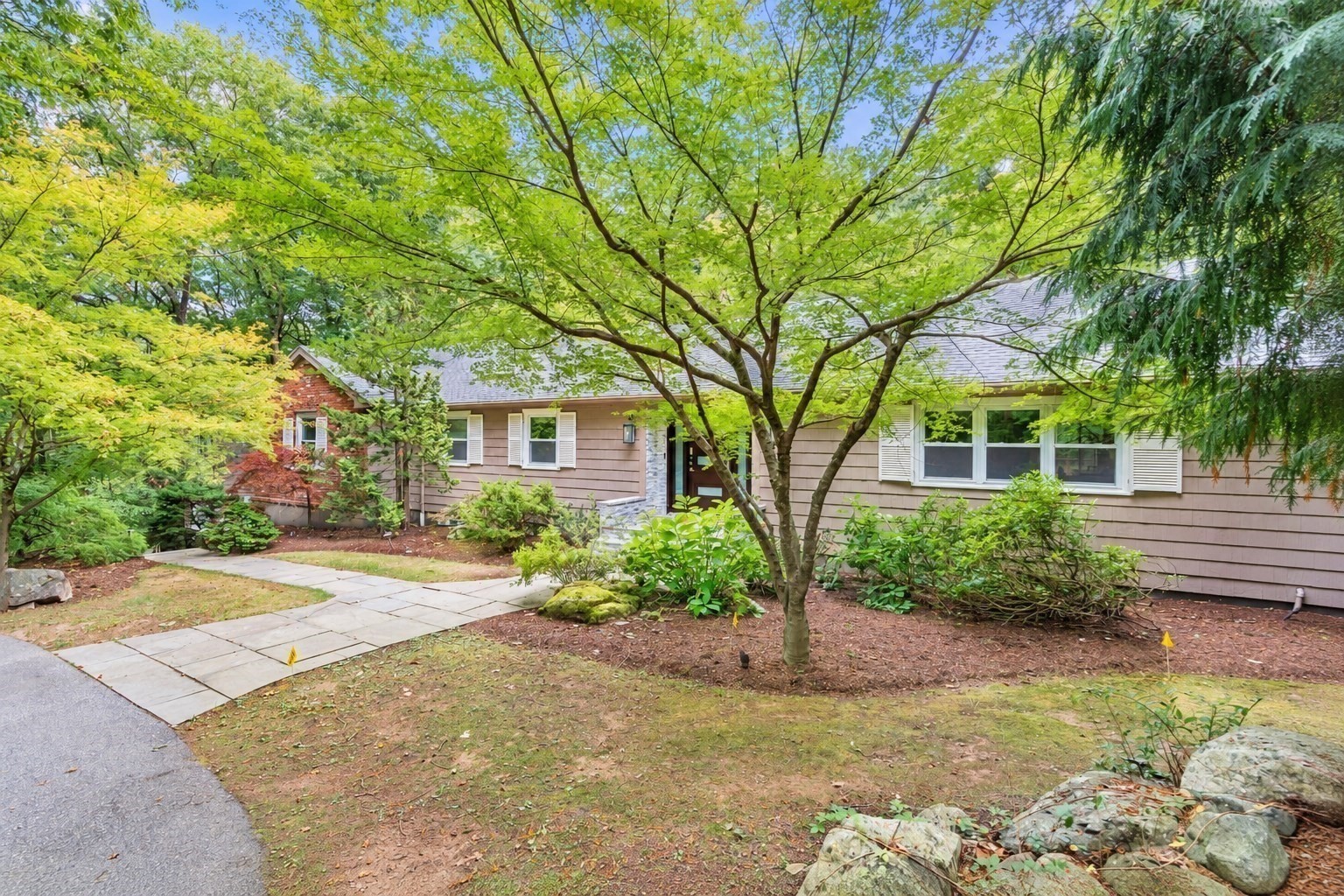 Single-story home with a brick entry, leafy trees, and a paved front walkway.