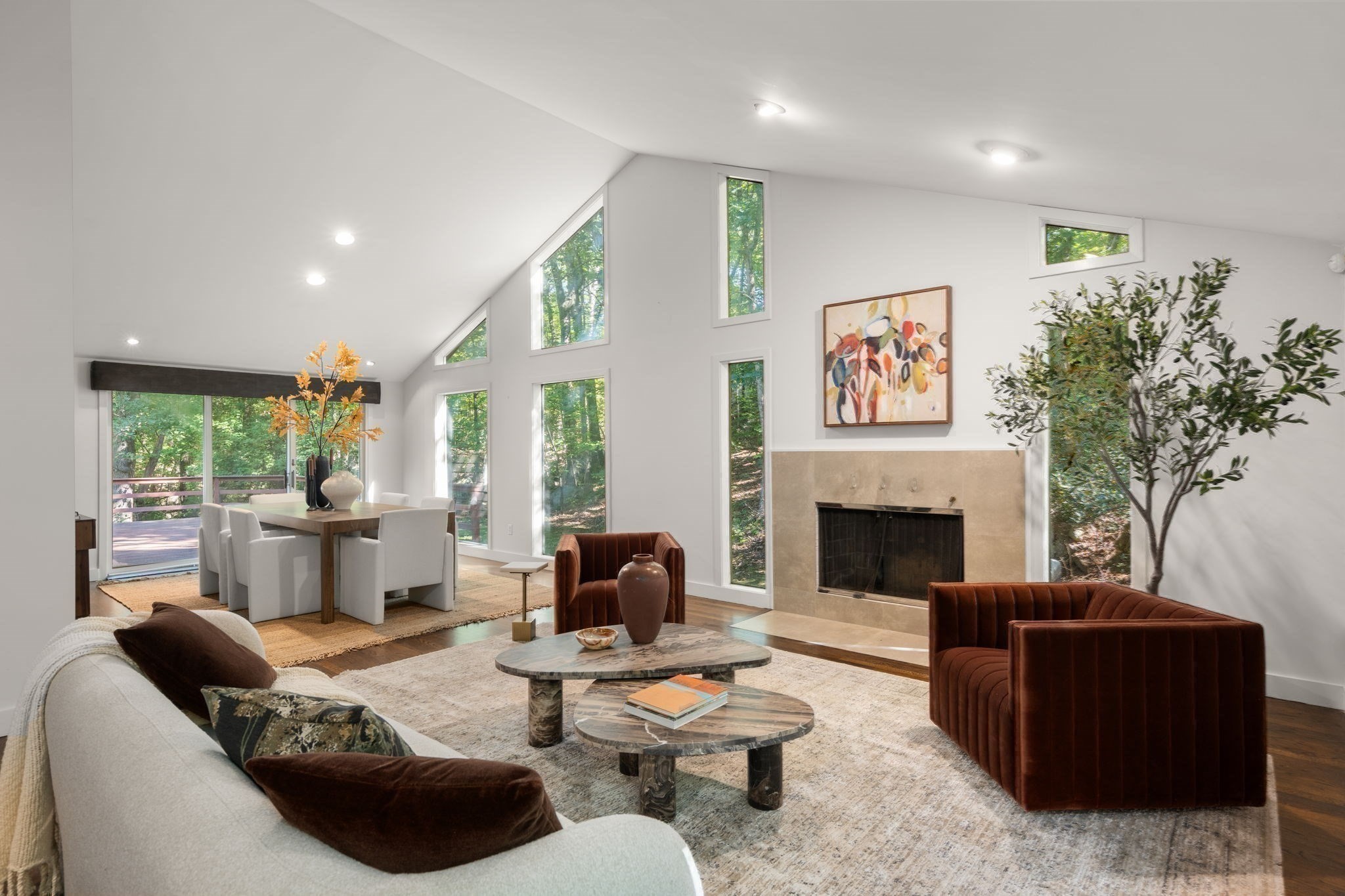 Vaulted living room with tall woodland-view windows, stone fireplace, and rust-colored armchairs.