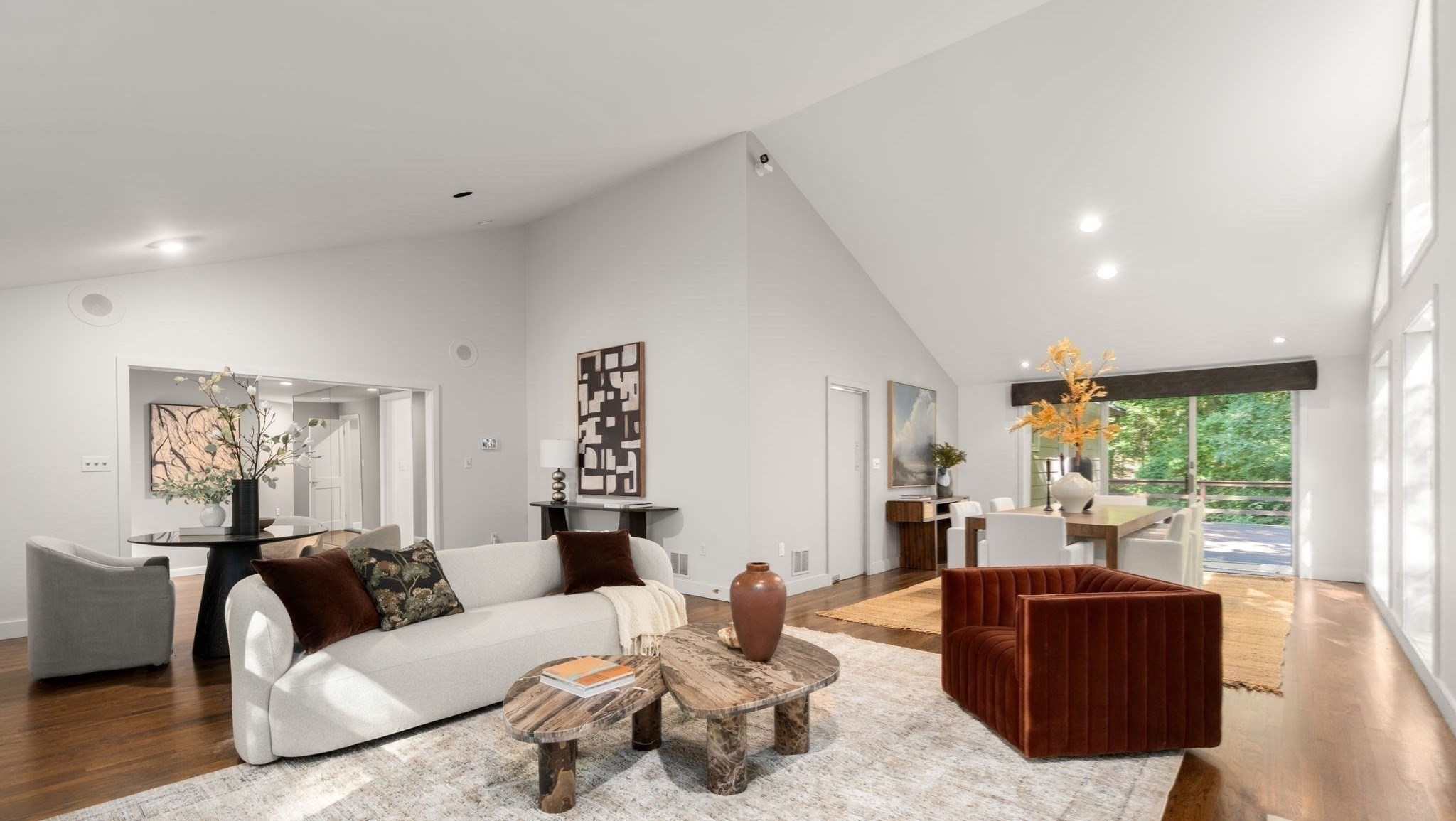 Open-plan living and dining room with a vaulted ceiling, white sofa, and large windows overlooking trees.